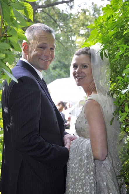 A man in a suit facing a woman in a wedding dress under a pergola.